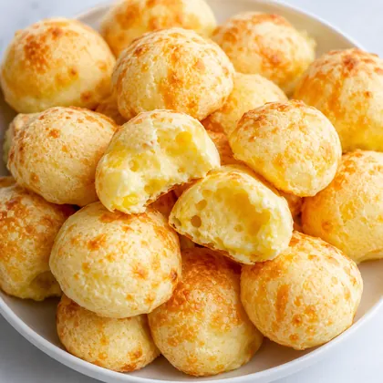 Pães de queijo dourados empilhados, um deles mordido mostrando o interior macio